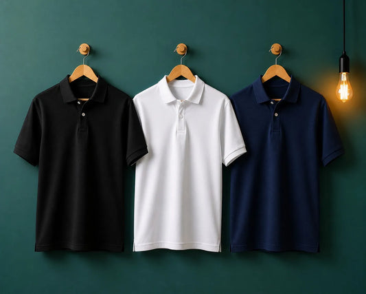 Three short-sleeve polo shirts in black, white, and navy blue hang side by side on wooden hangers against a dark green wall, with a warm hanging light bulb on the right.