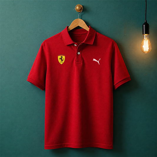 A red short-sleeve polo shirt hanging on a wooden hanger against a dark teal wall, with a yellow Ferrari shield logo on the left chest and a white Puma logo on the right chest. A glowing light bulb hangs to the right.