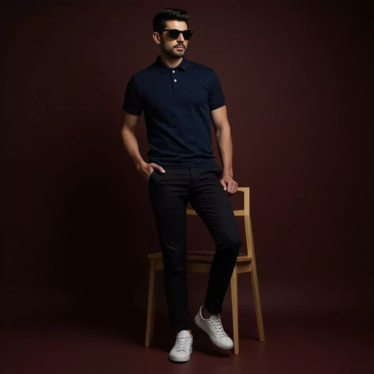 Man wearing a navy polo shirt, black pants, and white sneakers, sitting on a wooden stool against a dark background.