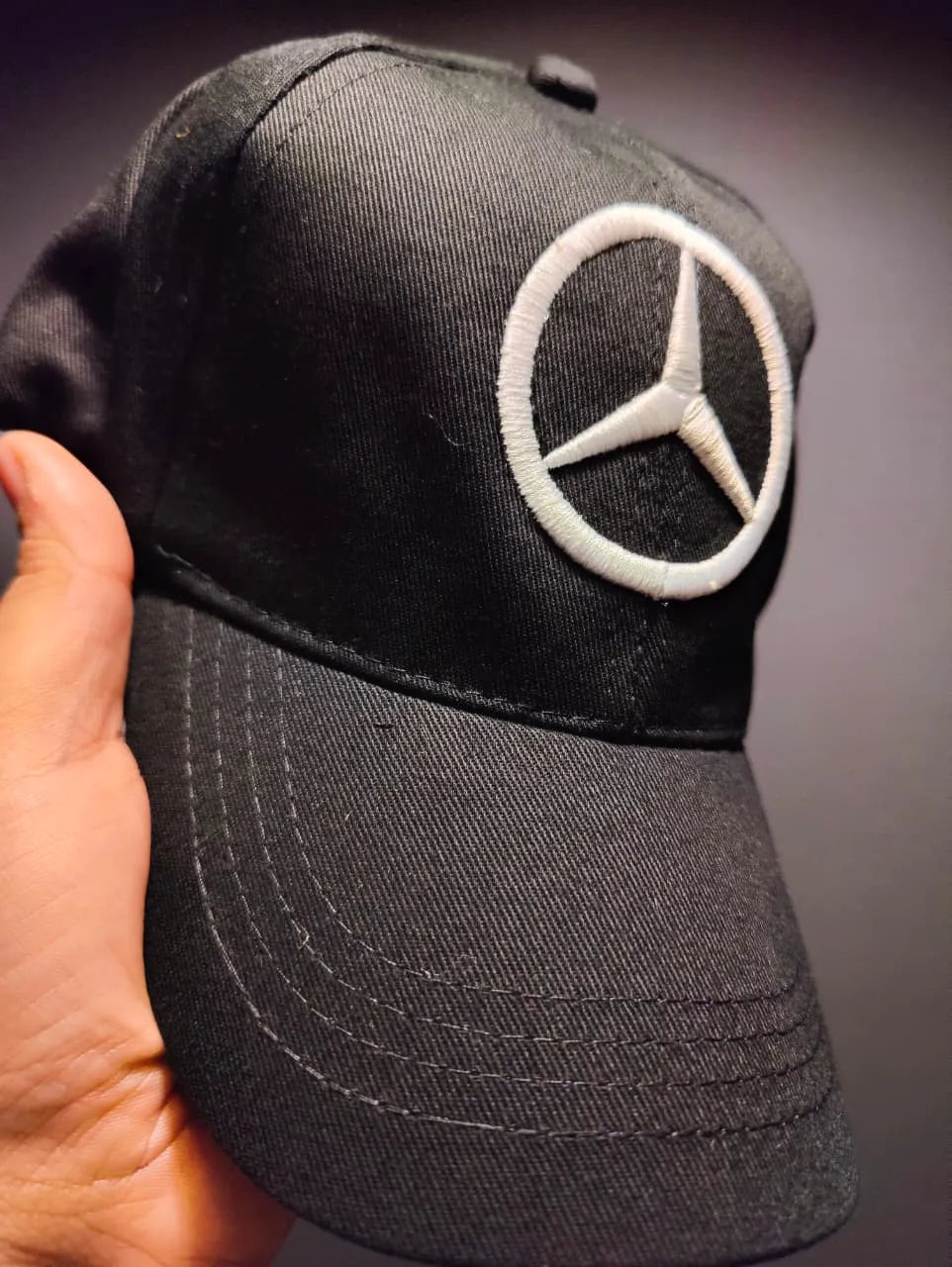 Black cap with Mercedes-Benz logo held by a hand against a dark background