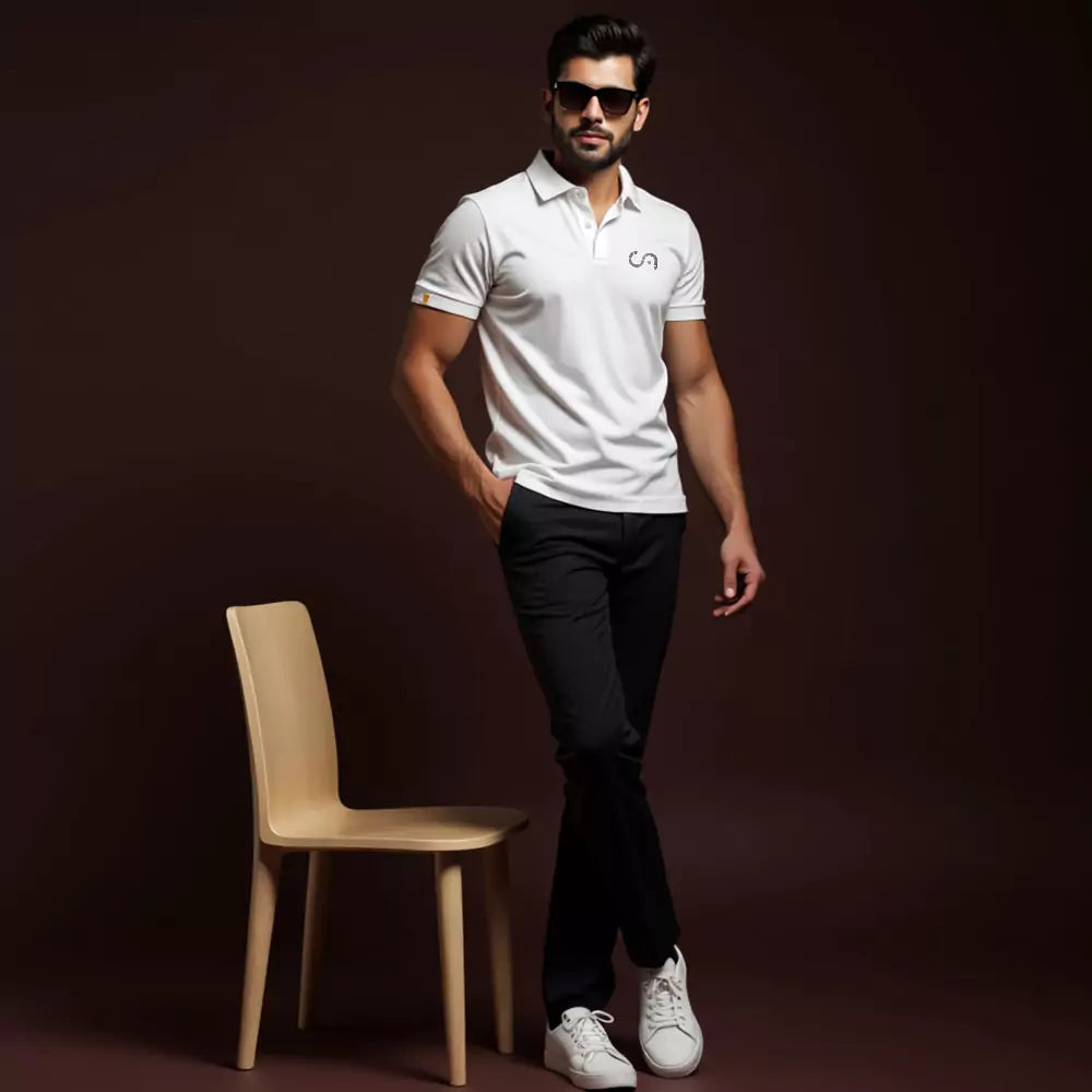 Man in a white polo shirt and black pants standing next to a wooden chair against a dark background